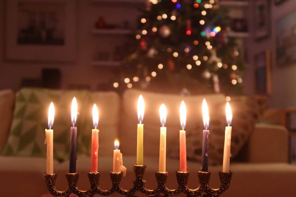 A photograph of a lit menorah with a Christmas tree in the background.