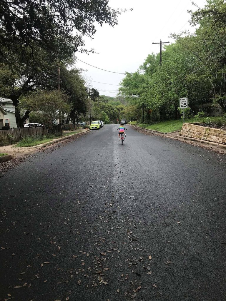 A photograph of a girl in a rainbow shirt, biking down a hill.