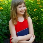 Emma Hoff, a girl with blond hair, wearing a red dress and holding a blue book.