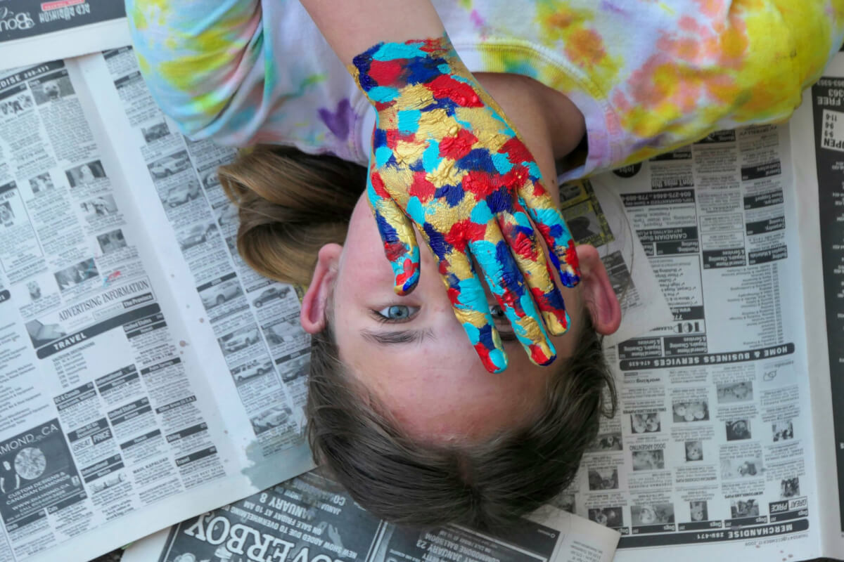 Colors by Sage Millen age 13. Paint-covered hand with visible eye against newspaper backdrop.